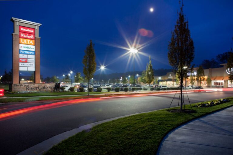 A nighttime view of a shopping center with illuminated signs and light trails from passing cars.